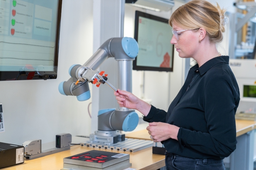 Zu sehen ist eine Frau, die einem Roboterarm einen Stift übergibt.