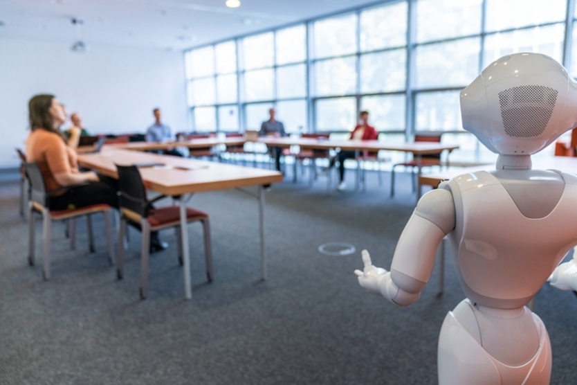 Zu sehen ist ein humanoider Roboter vor einer an einem Tisch sitzender Gruppe Menschen.