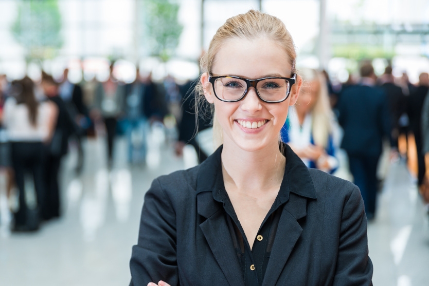 Eine junge blonde Frau im Business-Look mit dunkel umrandeter Brille steht mit selbstbewusst verschränkten Armen in einer Messehalle und lächelt den Betrachter:innen entgegen.